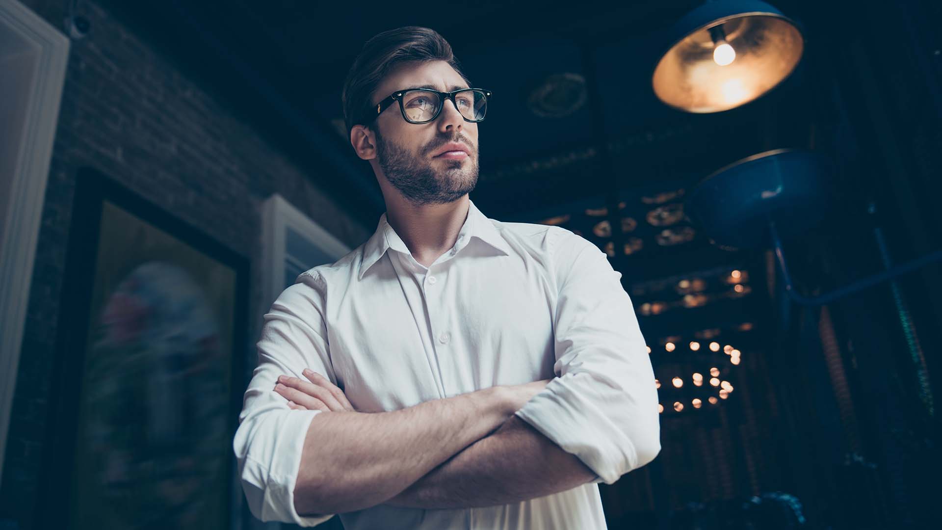 Brunette man in glasses and white shirt with his hands folded in front of him, looking ahead with a smolder.